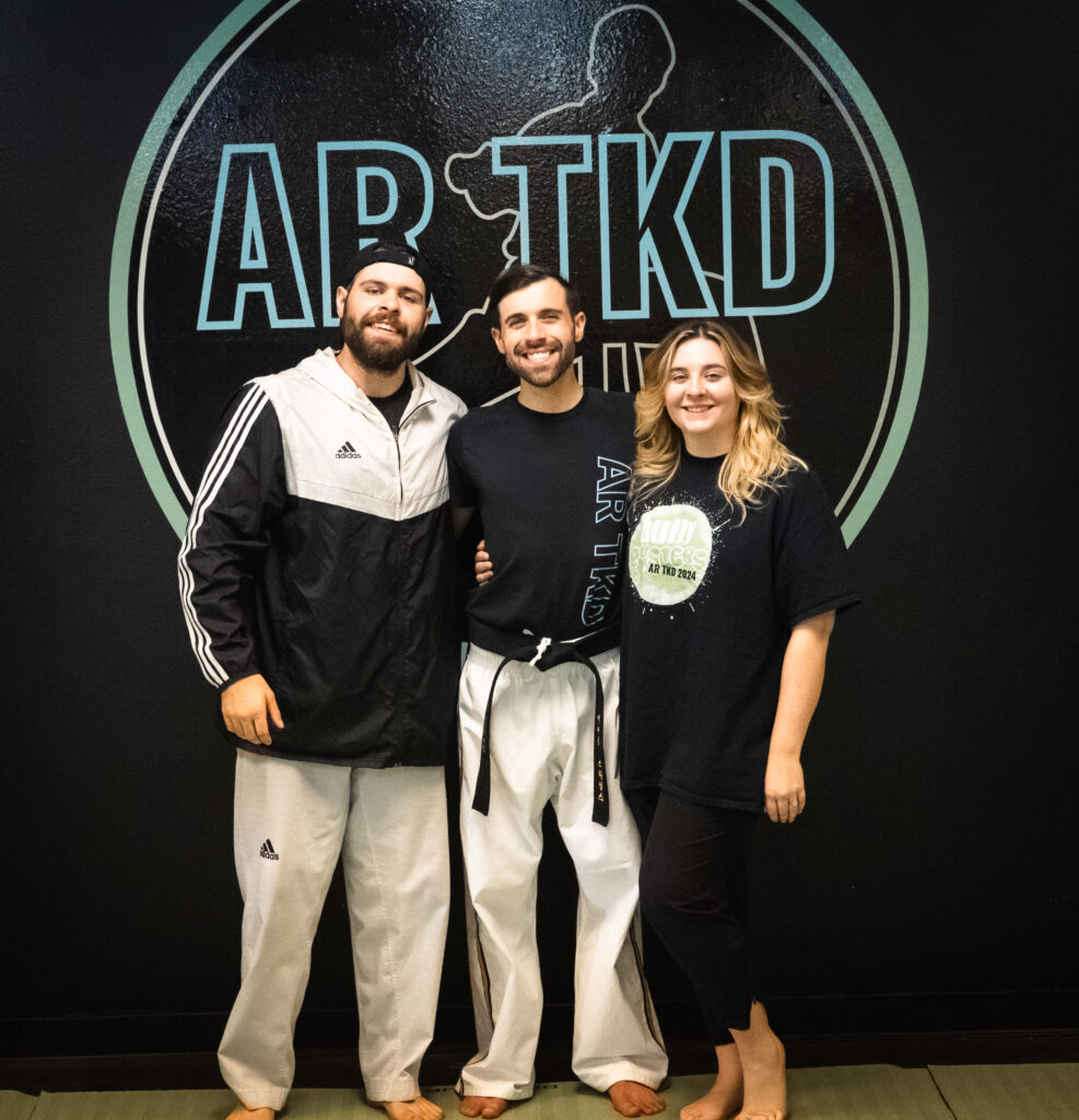 Family members and AR TKD instructors Andrew Stiles, AJ Stiles, and Alexis Gill standing together in front of the AR TKD logo, showcasing their unity and dedication to teaching.